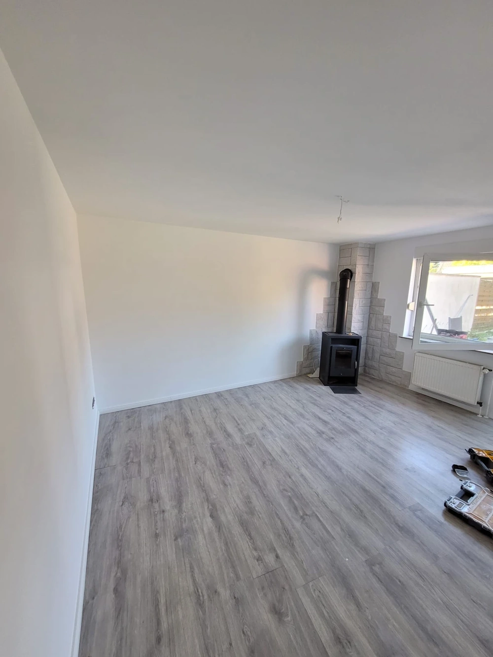 Laminat verlegen: Helle Wohnstube mit frisch verlegtem grauen Laminatboden, weiß gestrichenen Wänden und montiertem Kaminofen