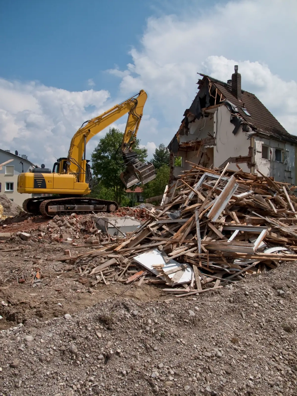 Gelber Bagger führt Abrissarbeiten an altem Wohnhaus durch, vor ihm liegen ein großer Trümmerhaufen und freigelegte Ziegel