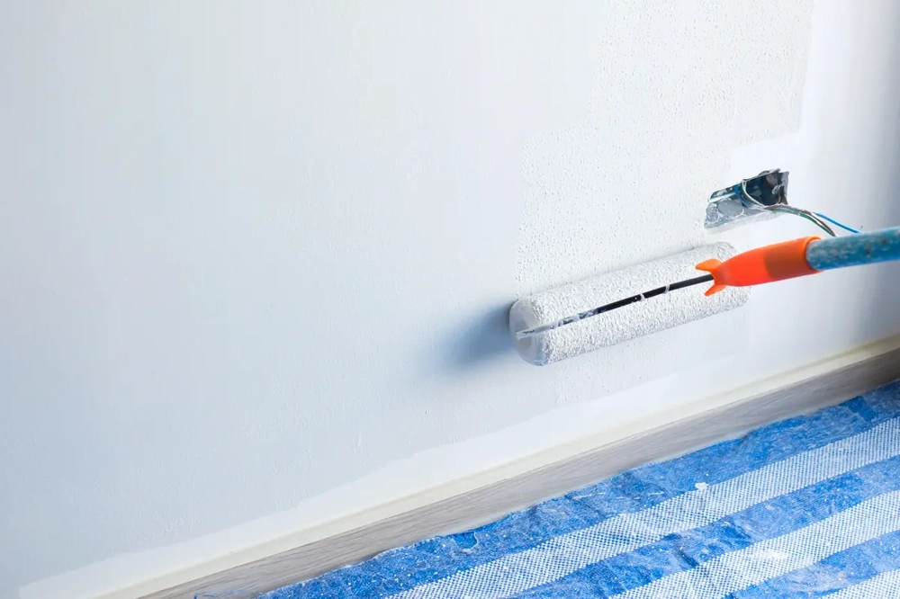 Malerarbeiten: Farbrolle streicht Wand in Weiß, Sockelleiste sichtbar, Boden mit Schutzfolie abgedeckt